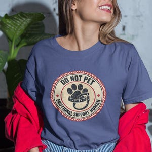 May include: A blue t-shirt with a circular graphic that reads "DO NOT PET EMOTIONAL SUPPORT HUMAN". The graphic features a paw print and a dog collar. The person is wearing a red jacket and blue striped jeans.