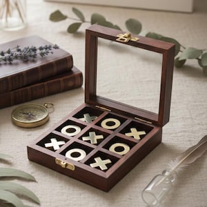 Op de afbeelding: Een houten tic-tac-toe spel met gouden en zilveren stukken. Het spel wordt getoond in een open, donkere houten doos met een glazen bovenkant. Andere items zijn boeken, een kompas en een kleine vaas met bloemen.