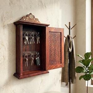 Könnte beinhalten: Ein dunkelbrauner Schlüsselhalter aus Holz mit dekorativer Oberseite und einer Tür mit Lochmuster. Der Schlüsselhalter ist an einer strukturierten Wand befestigt und hält mehrere Schlüssel. Ein modernes Wohnaccessoire.