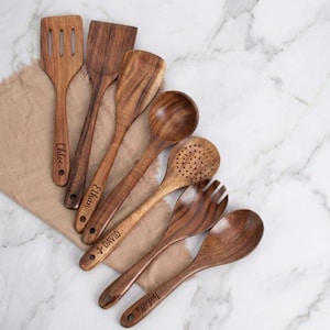 Könnte beinhalten: Eine Sammlung von sieben hölzernen Küchenutensilien, darunter Spatel, Löffel und eine Gabel. Jedes Utensil hat eine warme braune Farbe und einen personalisierten Namen auf dem Griff. Die Utensilien sind auf einem beigen Tuch vor einem weißen Marmorhintergrund angeordnet.