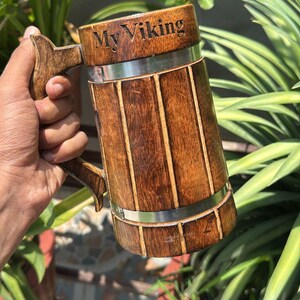 K&ouml;nnte beinhalten: Ein zylindrischer Bierkrug aus Holz mit vertikalen Lamellen, einem stabilen Griff und zwei horizontalen silberfarbenen Metallb&auml;ndern. Der obere Teil des Kruges ist mit dem Text My Viking in einer dunklen, klaren Schrift graviert.