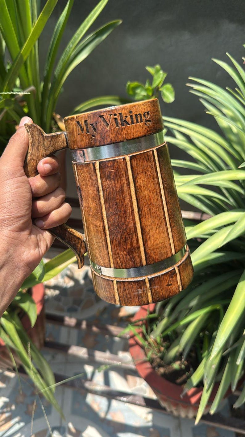 K&ouml;nnte beinhalten: Ein zylindrischer Bierkrug aus Holz mit vertikalen Lamellen, einem stabilen Griff und zwei horizontalen silberfarbenen Metallb&auml;ndern. Der obere Teil des Kruges ist mit dem Text My Viking in einer dunklen, klaren Schrift graviert.