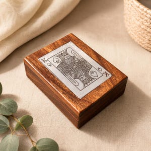 Puede incluir: Una caja rectangular de madera para naipes con un diseño plateado del Rey de Picas en la tapa. La caja es de madera marrón oscuro. El diseño de la carta es detallado e incluye el símbolo de picas.