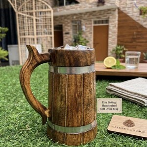 K&ouml;nnte beinhalten: Ein brauner Holzkrug mit geschwungenem Henkel und zwei silberfarbenen Metallb&auml;ndern. Der Krug enth&auml;lt Eisw&uuml;rfel. Ein kleines Schild daneben tr&auml;gt die Aufschrift: Etsy Listing | Handcrafted Soft Drink Mug. Ein h&ouml;lzerner Untersetzer ist mit einem Tannenzapfen und dem Text NATURE'S CHOICE graviert.