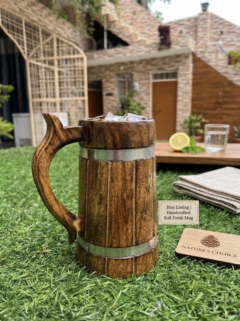 K&ouml;nnte beinhalten: Ein brauner Holzkrug mit geschwungenem Henkel und zwei silberfarbenen Metallb&auml;ndern. Der Krug enth&auml;lt Eisw&uuml;rfel. Ein kleines Schild daneben tr&auml;gt die Aufschrift: Etsy Listing | Handcrafted Soft Drink Mug. Ein h&ouml;lzerner Untersetzer ist mit einem Tannenzapfen und dem Text NATURE'S CHOICE graviert.