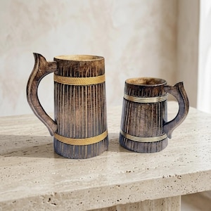 Könnte beinhalten: Zwei Bierkrüge aus Holz mit Griffen. Die Krüge sind dunkelbraun mit vertikalen Rillen und mit goldfarbenem Seil und Metallbändern umwickelt. Die Krüge sind unterschiedlich groß und stehen auf einer hellen Oberfläche.