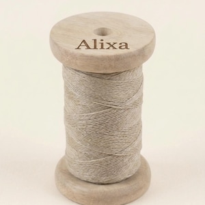 May include: A light brown wooden spool wrapped with beige thread. The top circular wooden flange features the name Alixa engraved in a dark brown serif font. The spool has a central hole and a smooth, natural wood grain finish throughout.