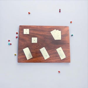 Puede incluir: Un tablero de juego de madera oscura con incrustaciones de latón y naipes. El tablero es rectangular. Cuatro naipes, con el símbolo de picas, están abanicados en el tablero. Pequeñas cuentas decorativas de colores están esparcidas alrededor del tablero.