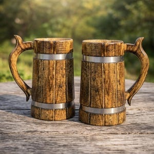 May include: Two wooden beer tankards with decorative handles and metal bands. The tankards are made of light brown wood and sit on a wooden surface. The background is a blurred view of green foliage.