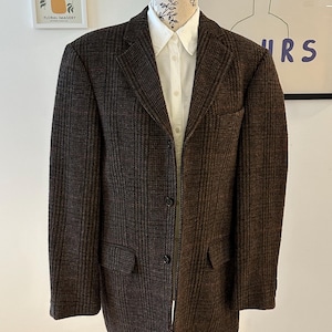 May include: A brown tweed blazer with a classic two-button fastening, displayed on a mannequin. The blazer features a notched lapel, two front pockets, and a white collared shirt underneath. The fabric has a subtle checkered pattern.
