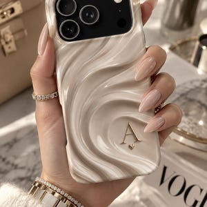 May include: A cream-colored phone case with a wavy, rippled texture and a glossy finish. The case features a black camera cutout area and a gold-colored letter A with a small heart symbol positioned below it on the lower right side of the case.