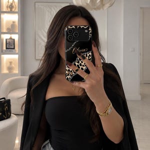 May include: A black smartphone case featuring a leopard print pattern, a diagonal silver glitter stripe, and the name Olivia written in gold script. The case has a camera cutout and covers the back of the phone, displaying a sleek design.