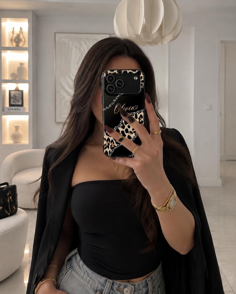 May include: A black smartphone case featuring a leopard print pattern, a diagonal silver glitter stripe, and the name Olivia written in gold script. The case has a camera cutout and covers the back of the phone, displaying a sleek design.
