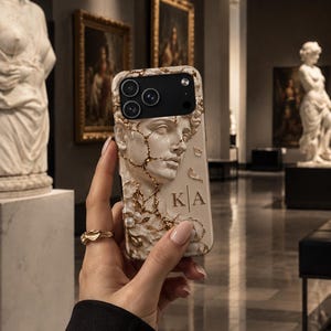 May include: A phone case featuring a raised, cream-colored relief of a classical bust with gold-toned cracks and accents. The design includes small white pearls and the letters K and A separated by a vertical line. The case has a black camera cutout.