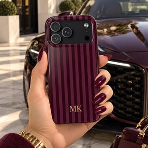 May include: A smartphone case featuring a deep burgundy and black vertical striped pattern with the gold-colored initials MK printed in the lower center. The case has a black camera module cutout with three circular lens openings and a flash.