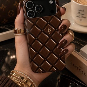Puede incluir: Una funda para smartphone de color marrón oscuro con un patrón de diamantes acolchado y brillante en la parte posterior. El centro de la funda muestra una letra B dorada con un pequeño símbolo de corazón debajo. El módulo de cámara es negro.