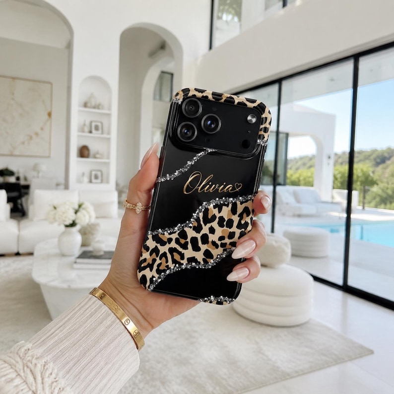 May include: A black smartphone case featuring a leopard print pattern, a diagonal silver glitter accent, and the name Olivia written in gold script. The case has a black camera module cutout and a leopard print border around the edges.