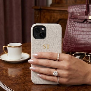 Könnte beinhalten: Eine hellbeige Handyhülle mit den goldenen Initialen "ST" wird in einer Hand gehalten. Die Hülle hat eine strukturierte Oberfläche. Die Hand ist mit Ringen geschmückt. Eine weiße Teetasse mit Goldrand steht auf einer Untertasse.