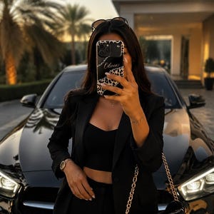 May include: A black smartphone case featuring a leopard print border and a central black panel with the name Olivia written in white script. The case has a camera cutout and is held up, obscuring the face of the individual holding the device.