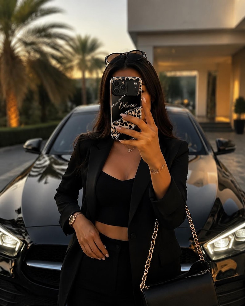 May include: A black smartphone case featuring a leopard print border and a central black panel with the name Olivia written in white script. The case has a camera cutout and is held up, obscuring the face of the individual holding the device.