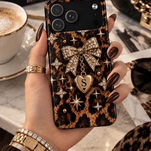 May include: A phone case featuring a brown and black leopard print pattern, adorned with a central gold-toned bow encrusted with clear crystals. A gold heart-shaped charm hangs from the bow, displaying the letter S in a black serif font.