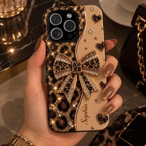May include: A smartphone case featuring a split design with a black and gold leopard print on one side and a solid beige colour on the other. A large, jewelled bow sits in the centre, accented by small heart-shaped crystals and the name Sophia.