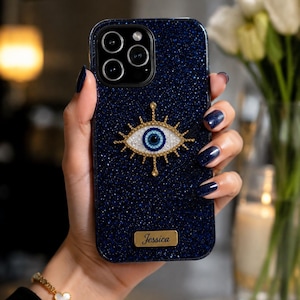 May include: A dark blue glittery phone case featuring a central gold-toned evil eye emblem with blue and white accents. The case includes a rectangular gold-toned nameplate at the bottom with the name Jessica engraved in a black script font.