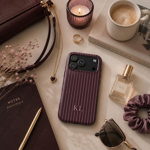 May include: A dark plum smartphone case with thin vertical white pinstripes and the monogram KL in white lettering. The case features a rectangular camera cutout with three circular lenses and a flash, centered among a collection of personal items.