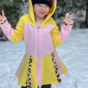 May include: A pink and yellow hooded sweatshirt with a leopard print trim. The sweatshirt has a zipper closure and a hood with a gray beanie.