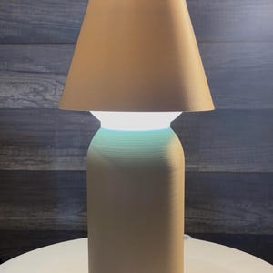 K&ouml;nnte beinhalten: Eine beige Tischlampe mit konischem Lampenschirm. Die Lampe hat einen zylindrischen Sockel und eine leuchtende wei&szlig;e Lichtquelle. Die Lampe steht auf einer wei&szlig;en Oberfl&auml;che vor einem dunklen Holzmaserungshintergrund.