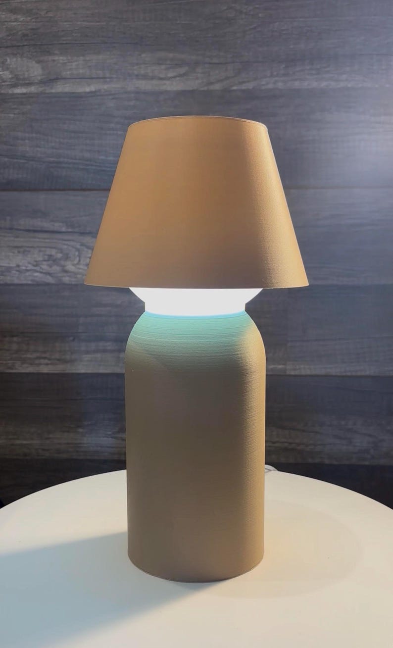 K&ouml;nnte beinhalten: Eine beige Tischlampe mit konischem Lampenschirm. Die Lampe hat einen zylindrischen Sockel und eine leuchtende wei&szlig;e Lichtquelle. Die Lampe steht auf einer wei&szlig;en Oberfl&auml;che vor einem dunklen Holzmaserungshintergrund.