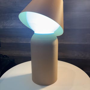 K&ouml;nnte beinhalten: Eine beige Tischlampe mit zylindrischem Sockel und konischem Lampenschirm. Der Lampenschirm ist abgewinkelt und gibt eine helle, leuchtende Lichtquelle frei. Die Lampe steht auf einer wei&szlig;en Oberfl&auml;che.