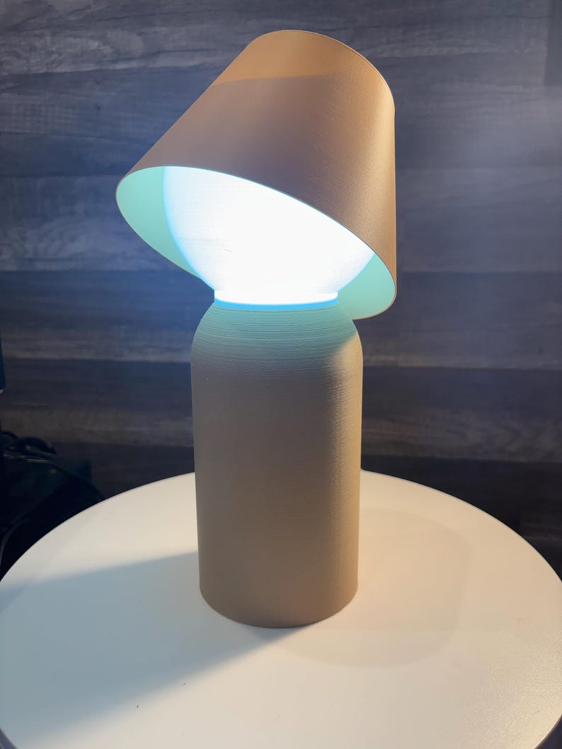 K&ouml;nnte beinhalten: Eine beige Tischlampe mit zylindrischem Sockel und konischem Lampenschirm. Der Lampenschirm ist abgewinkelt und gibt eine helle, leuchtende Lichtquelle frei. Die Lampe steht auf einer wei&szlig;en Oberfl&auml;che.