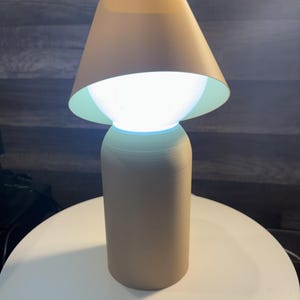 K&ouml;nnte beinhalten: Eine beige Tischlampe mit konischem Lampenschirm und zylindrischem Sockel. Die Lampe strahlt ein helles wei&szlig;es Licht aus, und die Innenseite des Lampenschirms ist hellgr&uuml;n. Die Lampe steht auf einer wei&szlig;en Oberfl&auml;che.