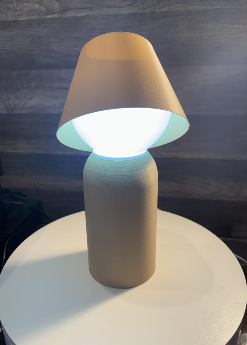 K&ouml;nnte beinhalten: Eine beige Tischlampe mit konischem Lampenschirm und zylindrischem Sockel. Die Lampe strahlt ein helles wei&szlig;es Licht aus, und die Innenseite des Lampenschirms ist hellgr&uuml;n. Die Lampe steht auf einer wei&szlig;en Oberfl&auml;che.