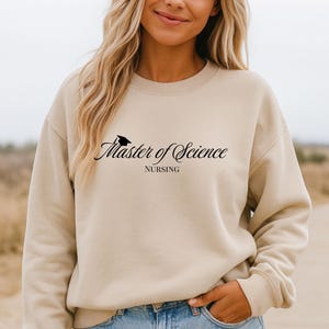 May include: A beige sweatshirt with the text "Master of Science" in a script font, with a graduation cap icon above the text. Below the text is the word "NURSING". The sweatshirt is a neutral color and has a crew neck.