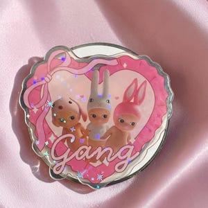 May include: A circular mirror featuring a heart-shaped frame with a pink ribbon design. Inside the heart are three small figures with rabbit ears, and the word Gang is written in cursive script at the bottom. The surface has a holographic finish.