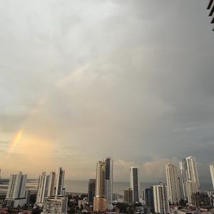 Op de afbeelding: Een stadsgezicht met talrijke hoge gebouwen onder een bewolkte hemel. Een gedeeltelijke regenboog boog over de lucht, wat een vleugje kleur aan de sc&egrave;ne toevoegt. De gebouwen vari&euml;ren in hoogte en ontwerp, met een watermassa in de verte.