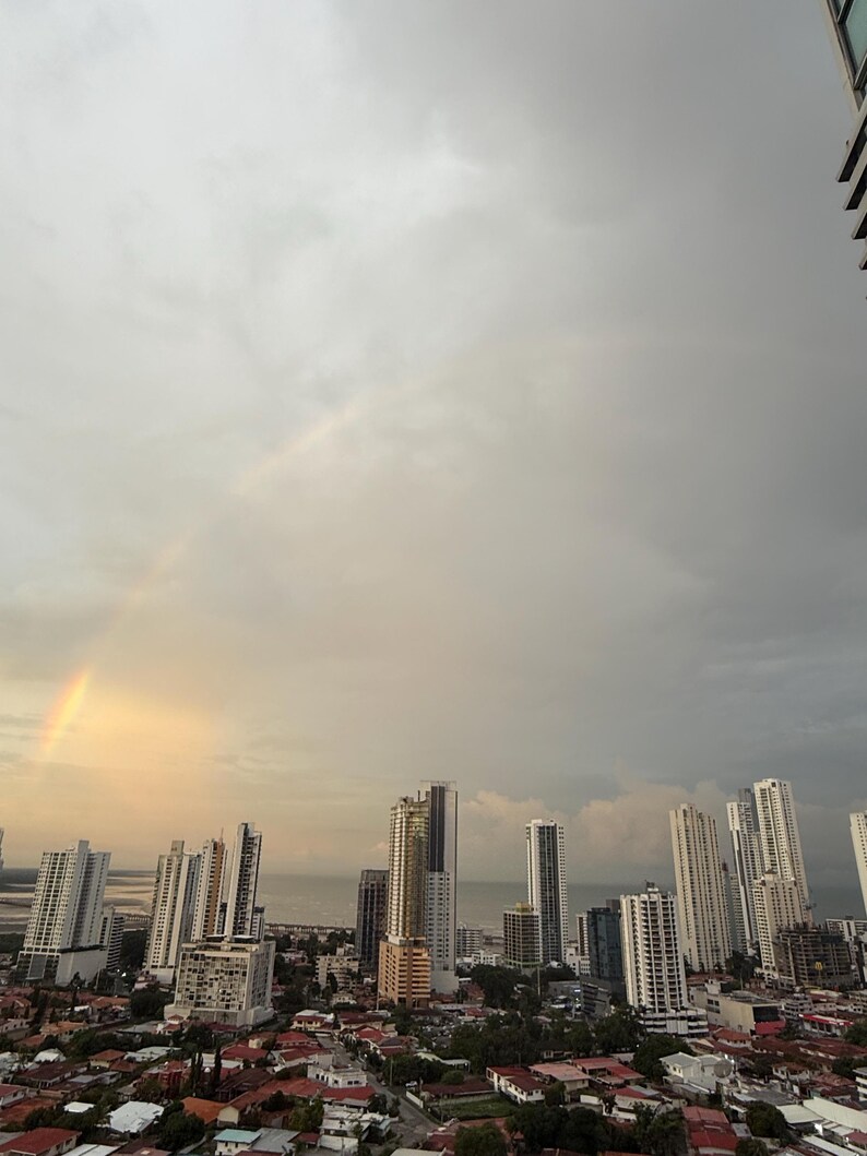 Op de afbeelding: Een stadsgezicht met talrijke hoge gebouwen onder een bewolkte hemel. Een gedeeltelijke regenboog boog over de lucht, wat een vleugje kleur aan de sc&egrave;ne toevoegt. De gebouwen vari&euml;ren in hoogte en ontwerp, met een watermassa in de verte.