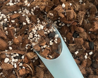 Könnte beinhalten: Nahaufnahme eines hellblauen Löffels, der teilweise mit einer Mischung aus brauner Kokosfaser, weißem Perlit und dunkelbraunen Stücken gefüllt ist. Die Blumenerde ist für Pflanzen und Gartenarbeit.
