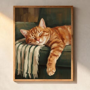 May include: A framed painting of a sleeping orange tabby cat. The cat is resting on a green couch with a striped blanket. The artwork is in a wooden frame and is displayed on a white wall.