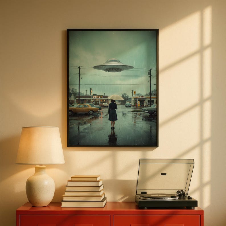 May include: Framed art print showing a person with an umbrella in a rainy street scene, with a diner and a UFO above. The artwork is in a black frame, with a vintage style and muted colours.