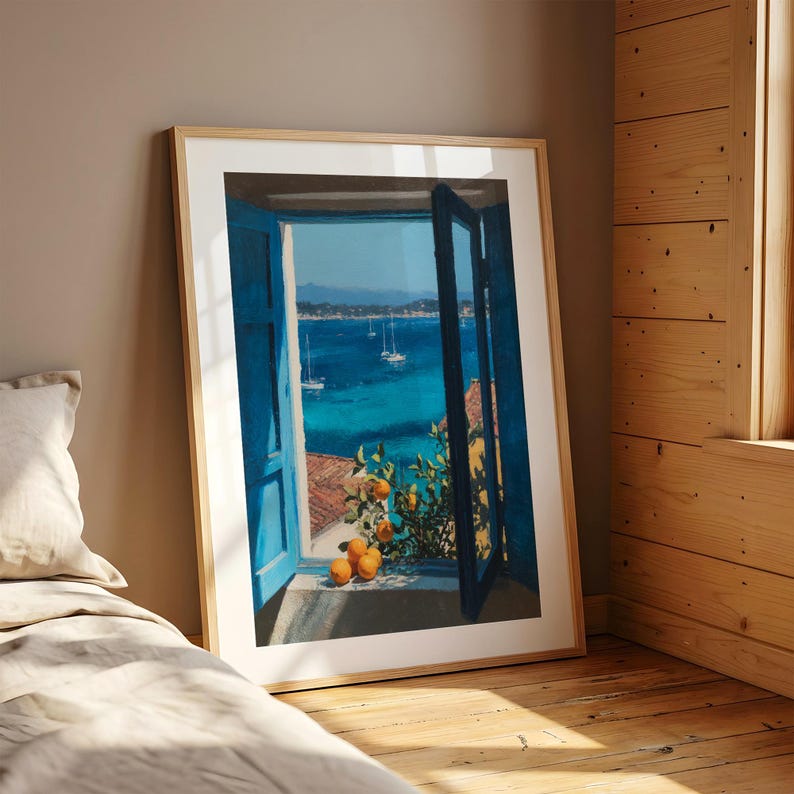 May include: A framed wall art print depicting an open window with bright blue shutters overlooking a vibrant turquoise sea with sailboats. In the foreground, a leafy orange tree branch with several ripe orange fruits rests on the window ledge.
