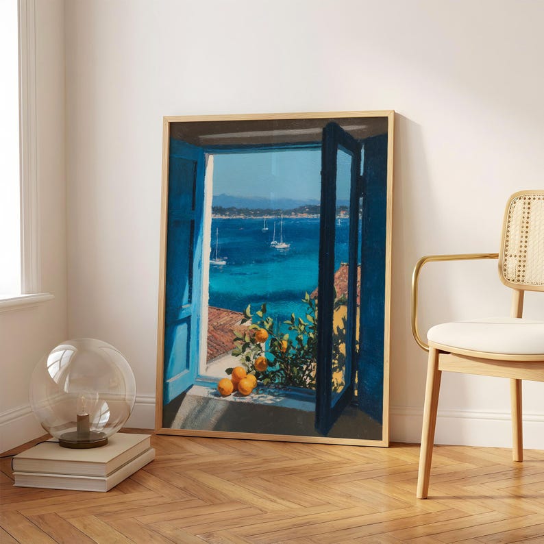 May include: A framed wall art print depicting an open window with bright blue shutters overlooking a deep blue sea with sailboats. In the foreground, an orange tree branch with ripe fruit rests on the windowsill, casting shadows in the sunlight.