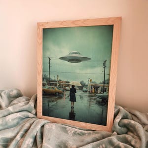 May include: A framed print shows a rainy street scene with a vintage diner and petrol station. A person with an umbrella stands in the rain, looking toward a hovering UFO. The print is in a light wooden frame.