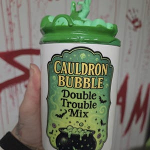 Könnte beinhalten: Ein weißer Keramikbehälter mit grünem Deckel und grüner handförmiger Dekoration. Der Behälter hat ein Etikett mit der Aufschrift "Cauldron Bubble Double Trouble Mix" mit einer Kesselgrafik, Fledermäusen und Sternen.