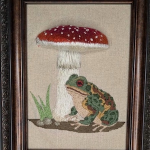 Op de afbeelding: Geborduurd kunstwerk in een donkerhouten lijst. De afbeelding toont een rood-witte paddenstoel met een groen-bruine kikker. Groen gras staat links van de paddenstoel. De achtergrond is een beige stof.