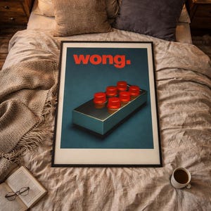 May include: A framed art print featuring the word "wong" in red letters above a teal and silver box with six red circular objects. The print is displayed on a bed with neutral-toned bedding, a book, and a cup of coffee.