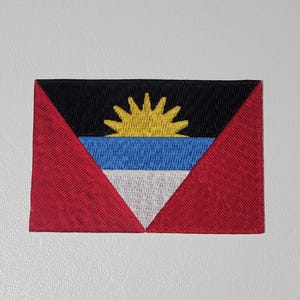 May include: Embroidered patch featuring the flag of Antigua and Barbuda. The flag design includes a black top, red sides, a blue and white stripe, and a yellow rising sun in the center. The patch is rectangular.