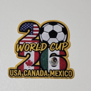 May include: A patch featuring the text "26 WORLD CUP USA CANADA MEXICO" in yellow. The number "26" is formed by the flags of the United States and Canada. A soccer ball sits atop the number, and the flag of Mexico forms the bottom of the "6".
